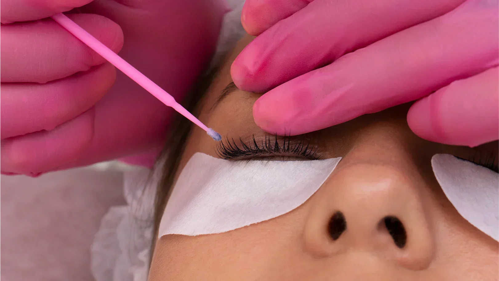 Une femme reçoit un réhaussement de cils, les yeux fermés, pendant qu'une technicienne porte des gants roses.