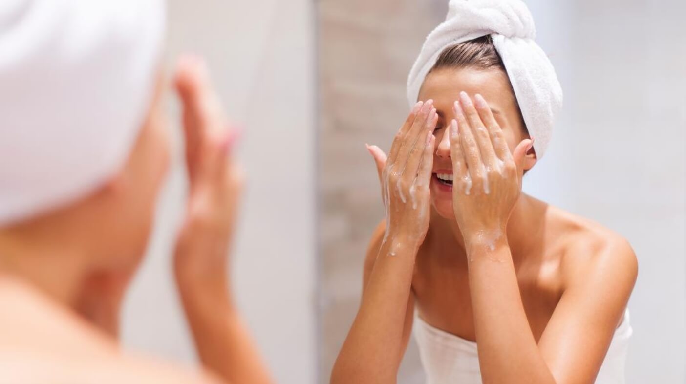 Une femme en serviette se lave le visage devant un miroir, illustrant des conseils pour une peau éclatante.