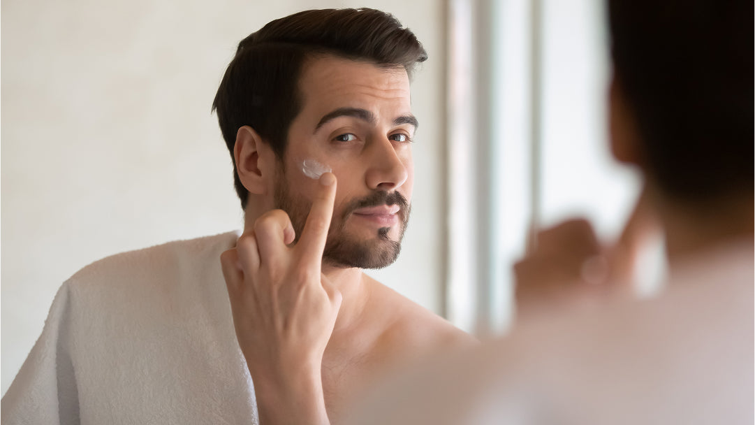 Un homme se regarde dans le miroir en appliquant de la crème hydratante sur son visage.